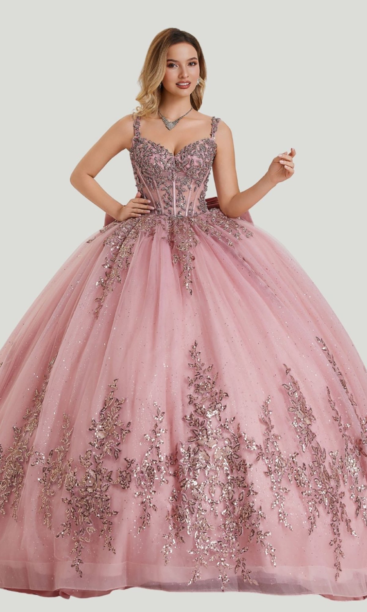 A woman wears the Quinceanera Dress 26406 by Stella Couture in mauve, featuring silver floral embellishments, glitter tulle spaghetti straps, and a fitted bodice, as she poses against a plain light background.